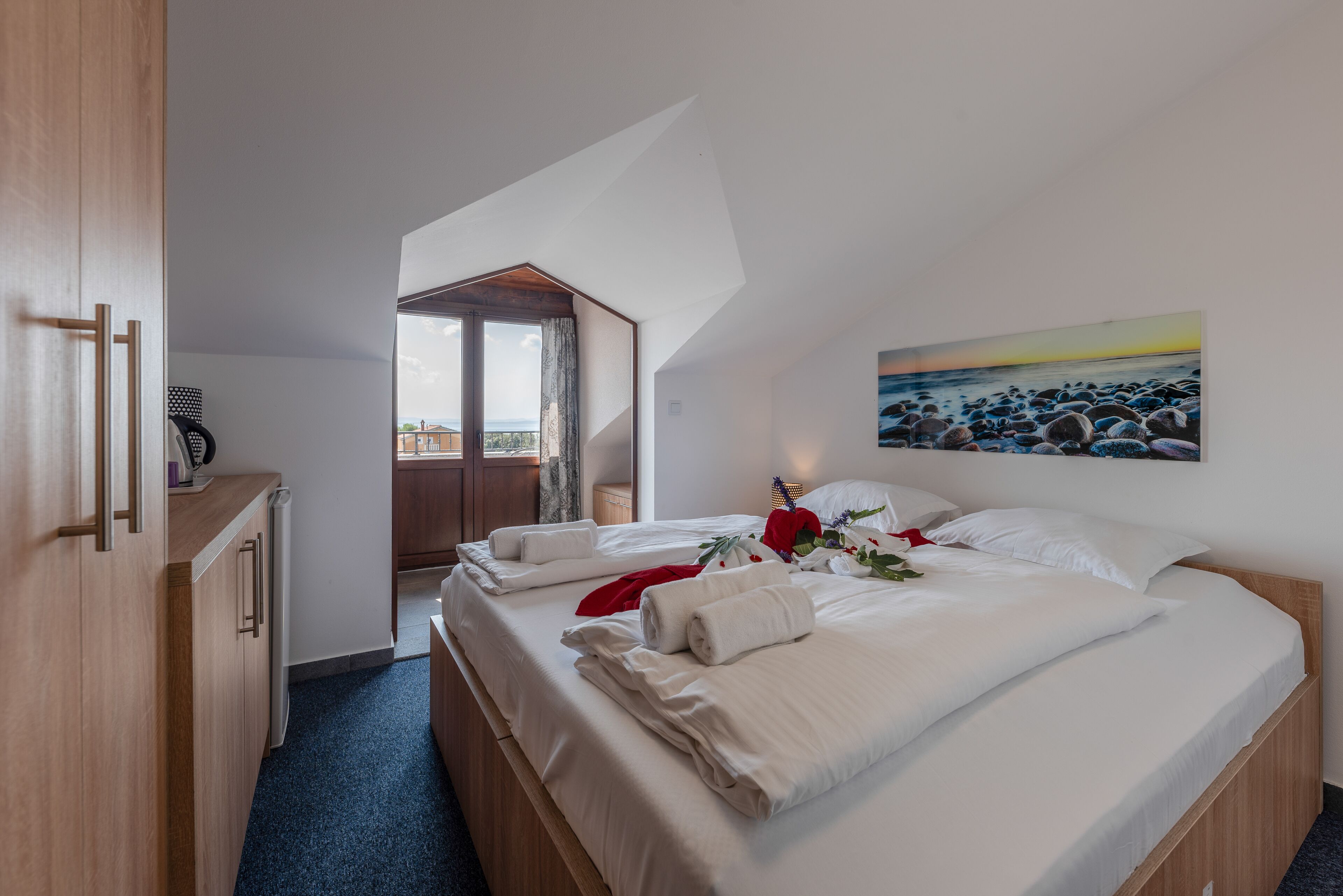 A bedroom with a large bed, a wooden wardrobe, a painting of a beach, and a view of the outdoors.