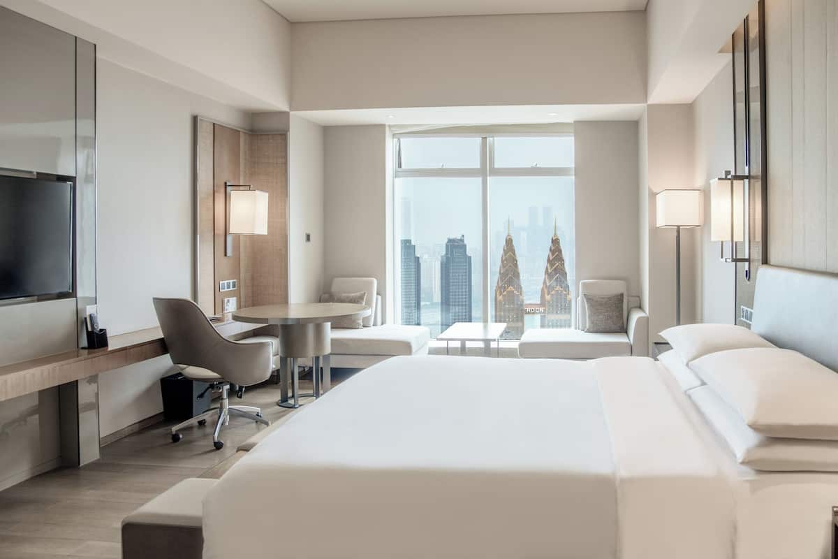 A modern hotel room with a large bed, a desk, two chairs, a small table, and a TV. The room has large windows with a city view.