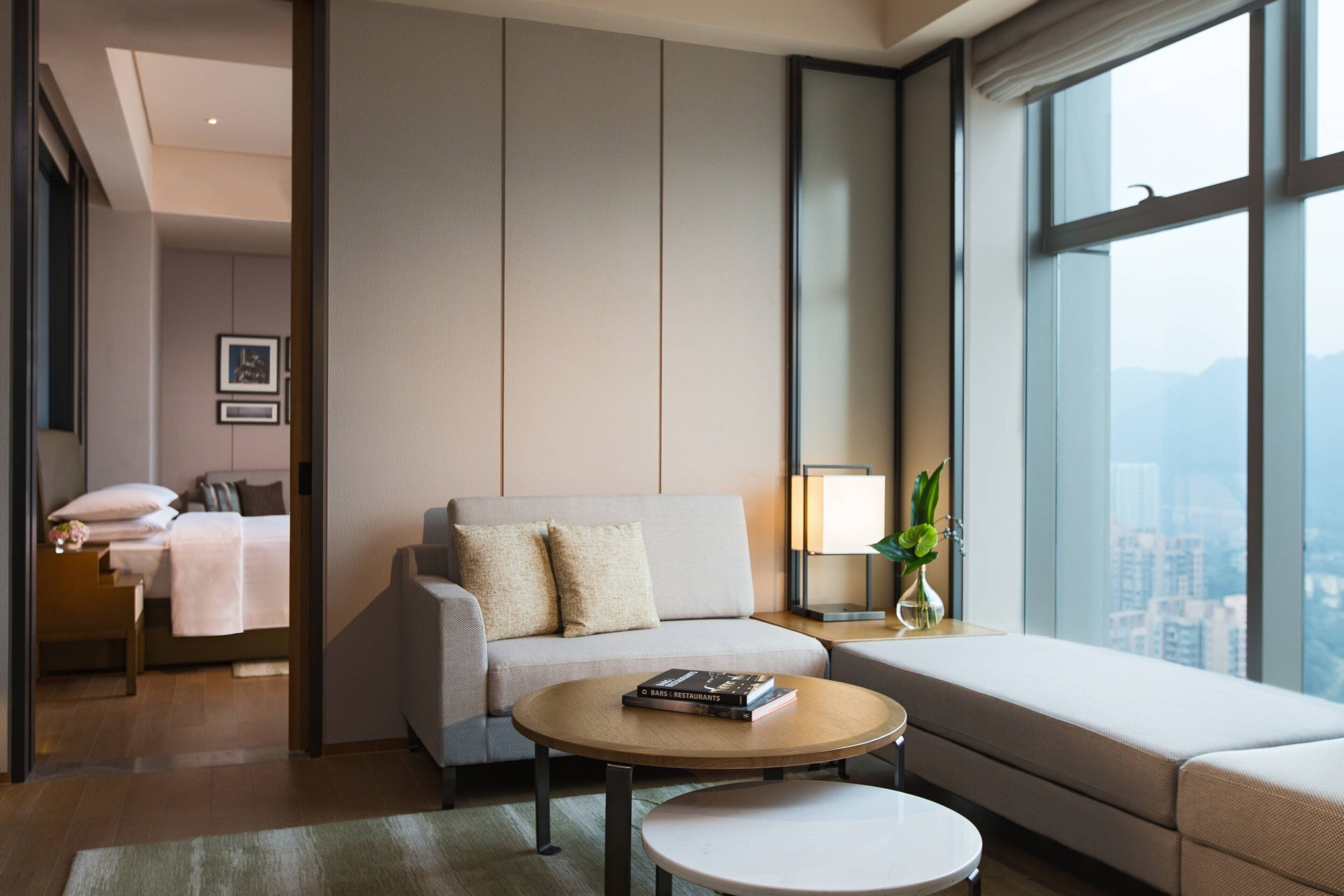 Una habitación de hotel moderna con sofá, una mesita redonda, una mesita de noche con lámpara y vistas a la ciudad a través de amplios ventanales.