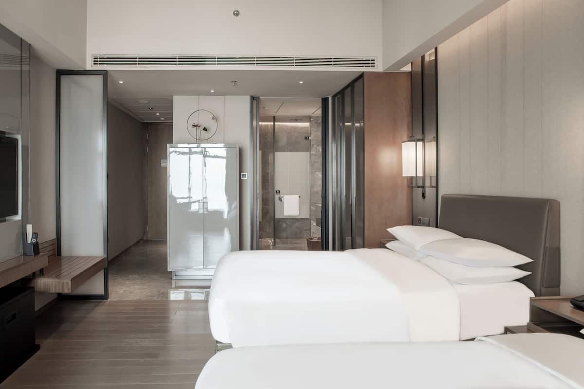 A modern hotel room with a large bed, a desk, and a television. There is a bathroom visible through a glass door.
