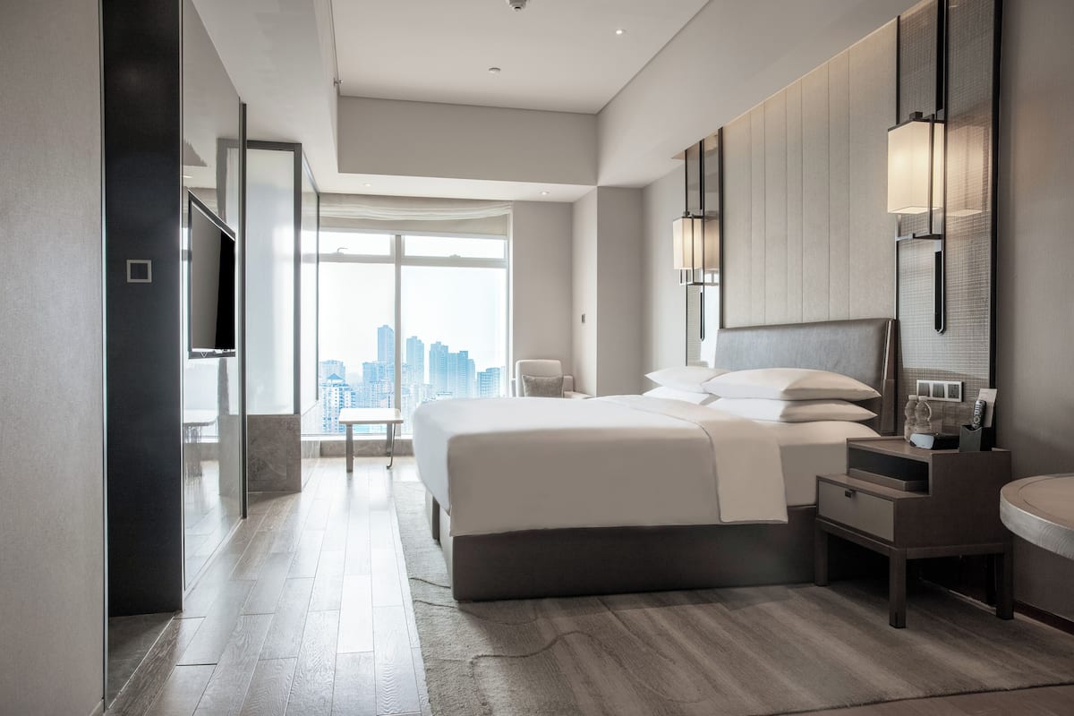 A modern hotel room with a large bed, a bedside table, a chair, and a view of the city.