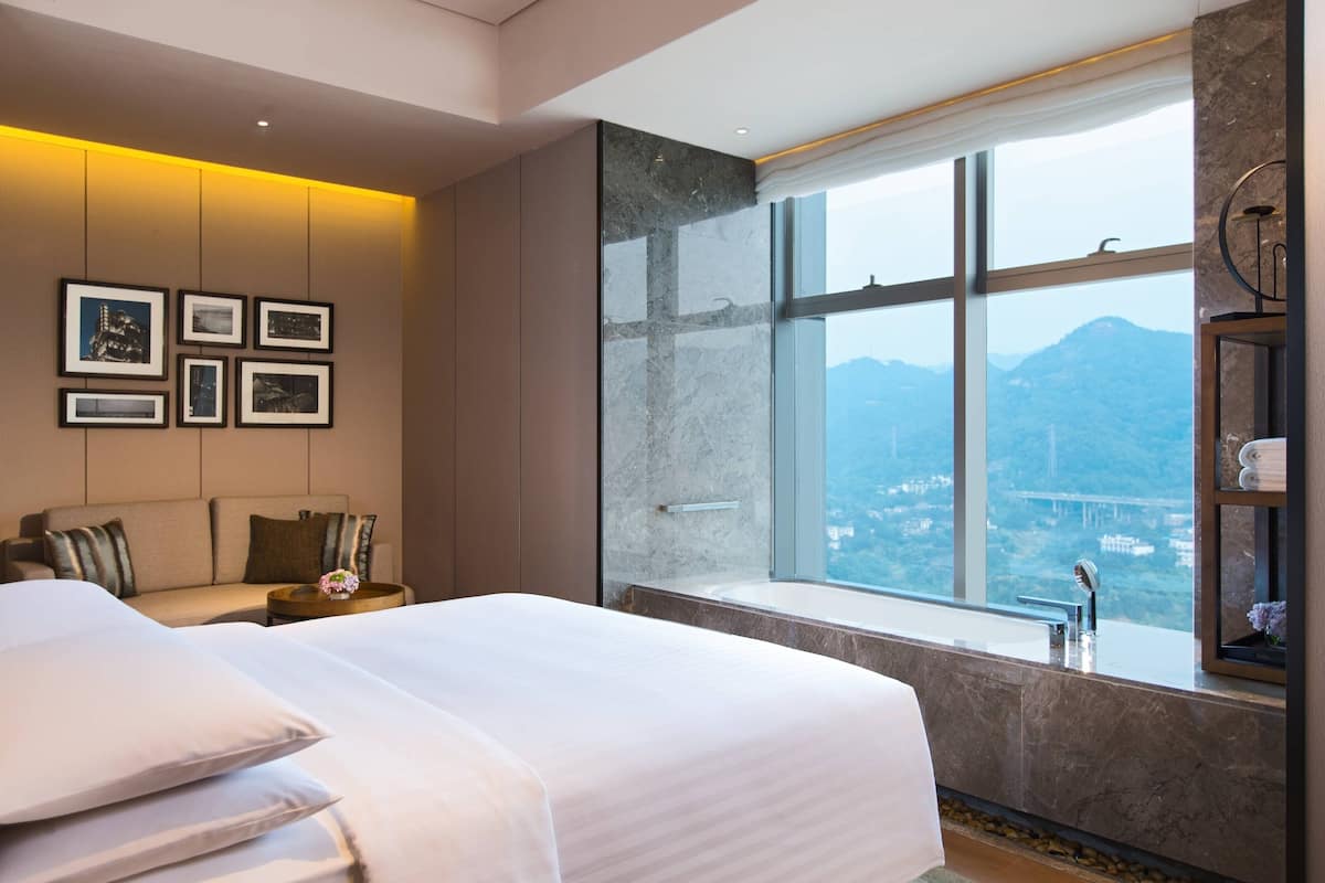 A modern hotel room with a large bed, a sofa, a small table with a vase, a large window with a city view, and framed pictures on the wall.
