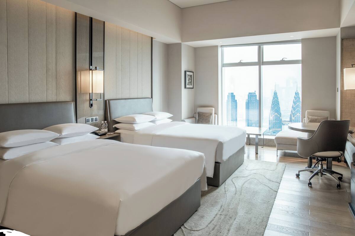 A hotel room with two beds, a desk, a chair, and a large window with a city view.