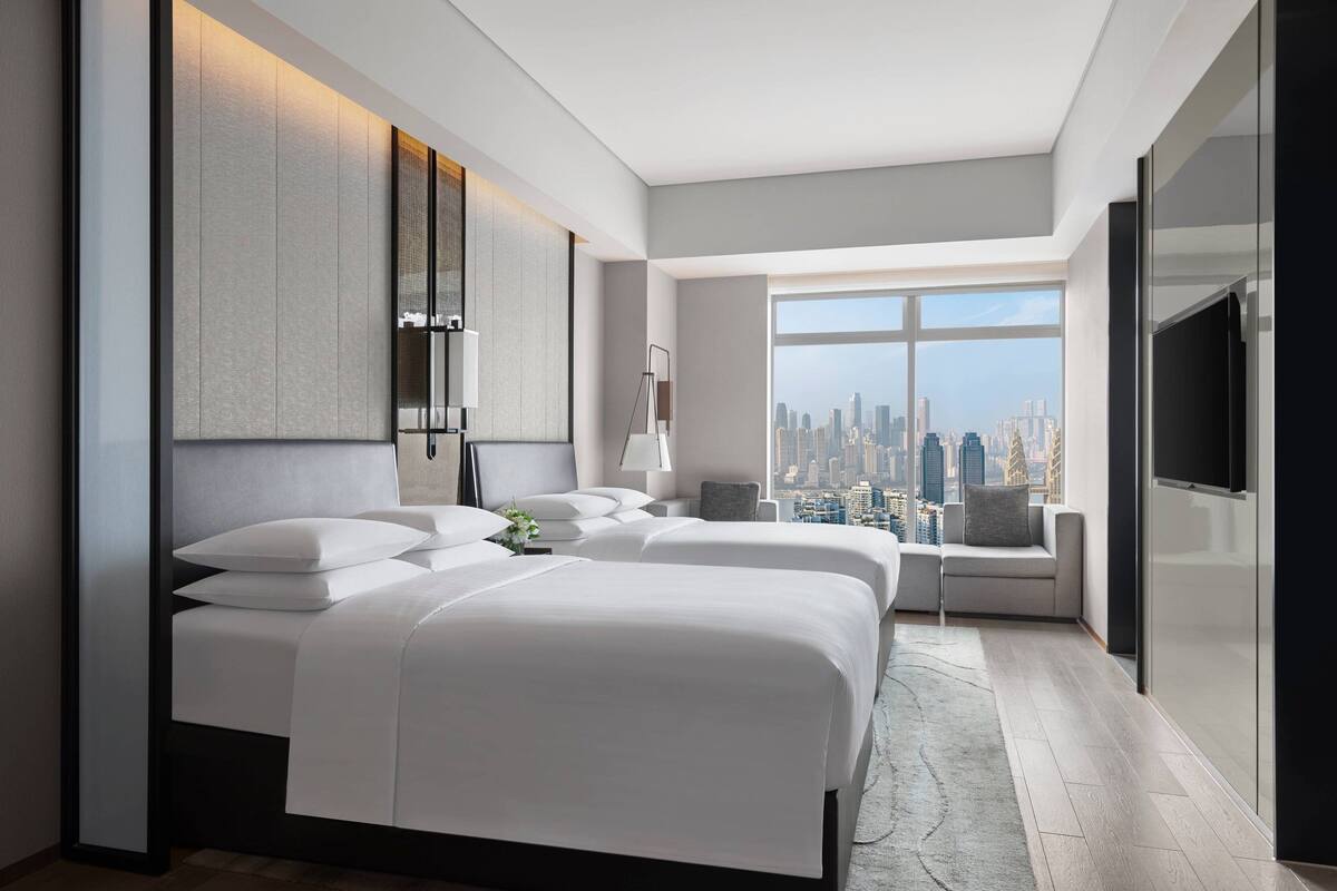 A modern hotel room with two large beds, a city view, a flat-screen TV, and a seating area with a cityscape view.