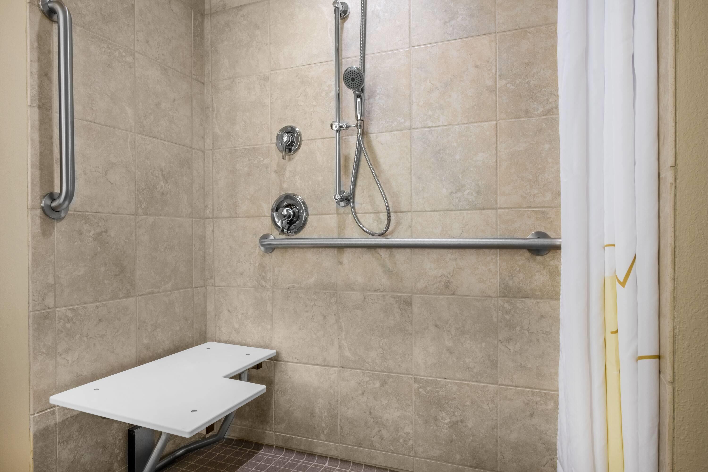 Un área de ducha con barra de apoyo, cabezal de ducha de mano y un banco blanco.