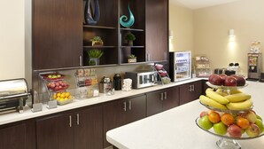 A kitchen area with a buffet setup including fruits, a coffee station, and a microwave.