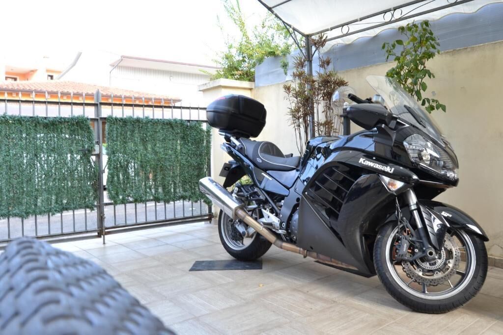 Une moto est garée sur une terrasse carrelée, avec une clôture en métal et de la verdure en arrière-plan.
