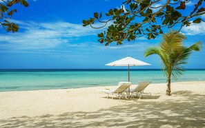 Una playa con dos sillas de playa blancas bajo una sombrilla blanca, una palmera y agua azul cristalina.