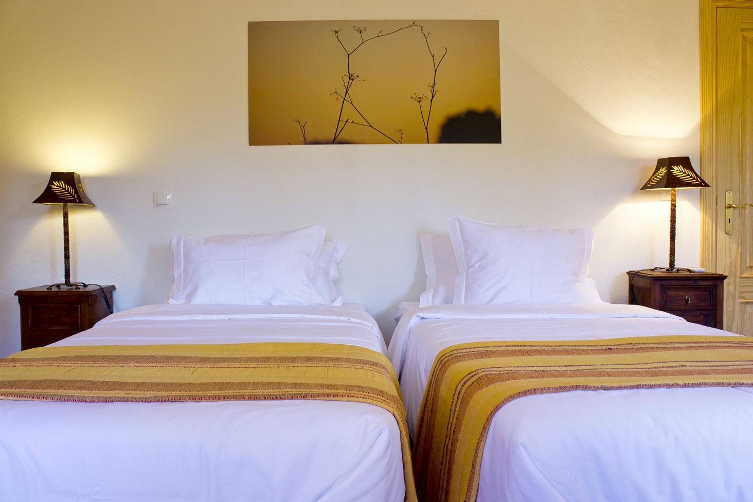 Habitación doble con dos camas, cada una con sábanas blancas y colchas a rayas. Hay dos mesitas de noche con lámparas, y un cuadro cuelga en la pared sobre las camas.