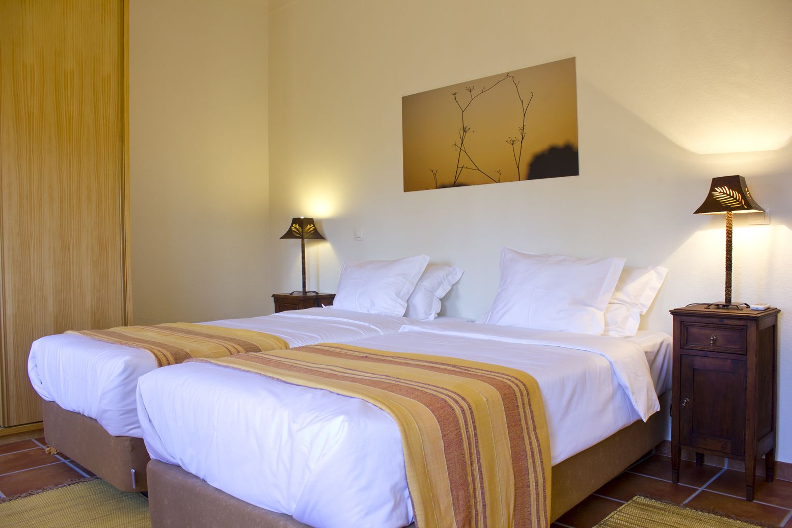 Una cama doble con ropa de cama blanca y una manta a rayas, dos mesitas de noche con lámparas, una obra de arte colgada en la pared y una mesita de noche de madera.