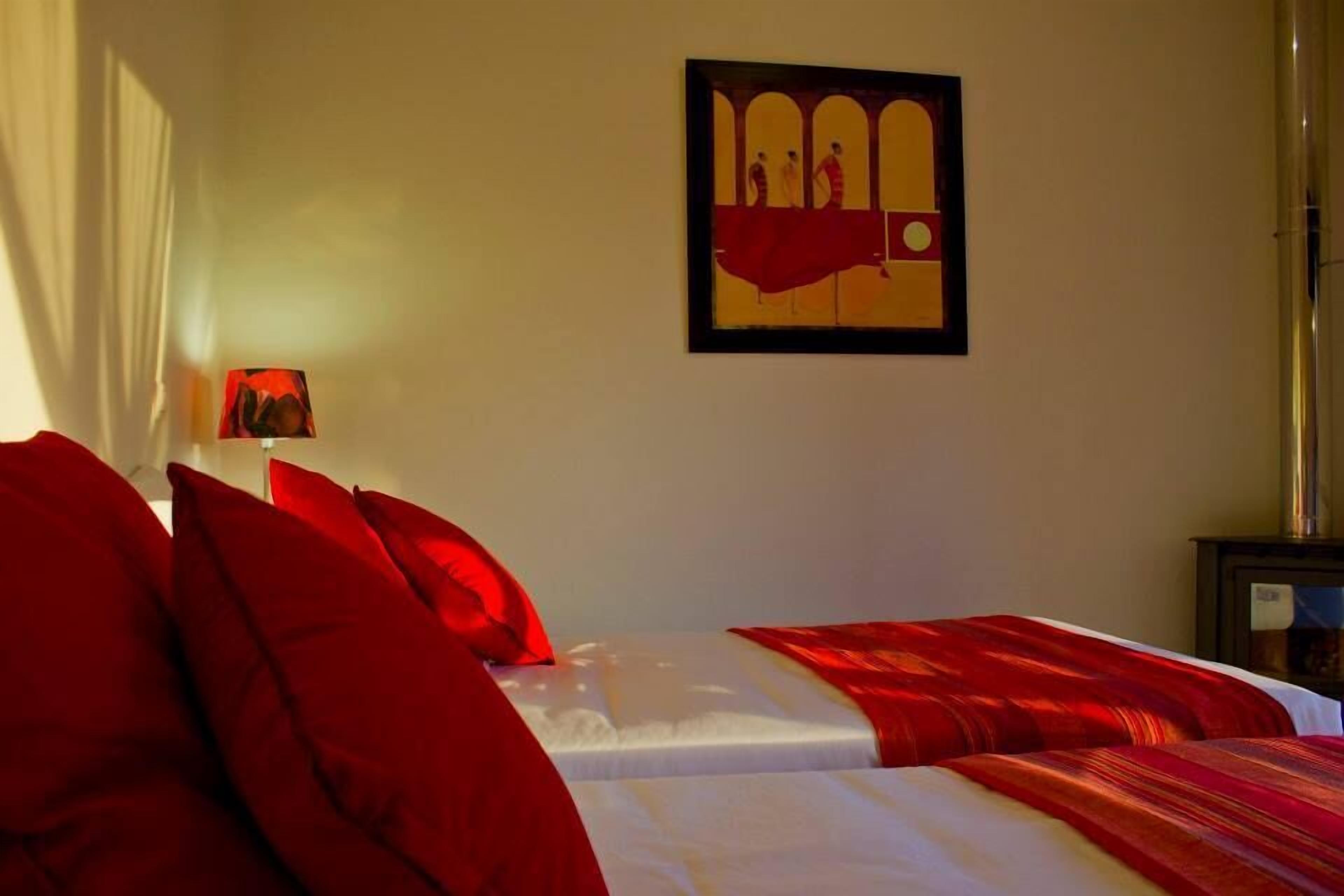 A hotel room with two beds, red bedding, a wall-mounted artwork, and a nightstand with a lamp.