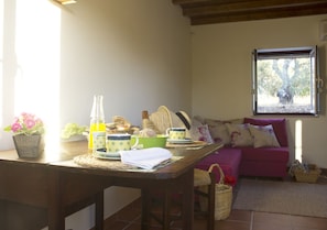 Un salón acogedor con una mesa de madera dispuesta para el desayuno, un sofá con cojines y una ventana con vistas a árboles.