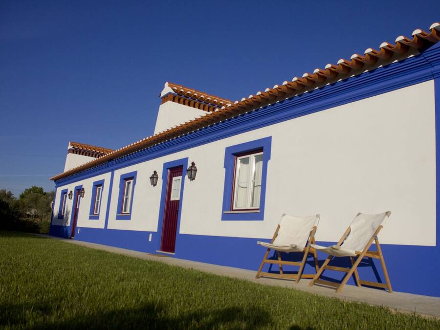 A two-story building with a blue and white facade, a red door, and a tiled roof. There are two white folding chairs outside.