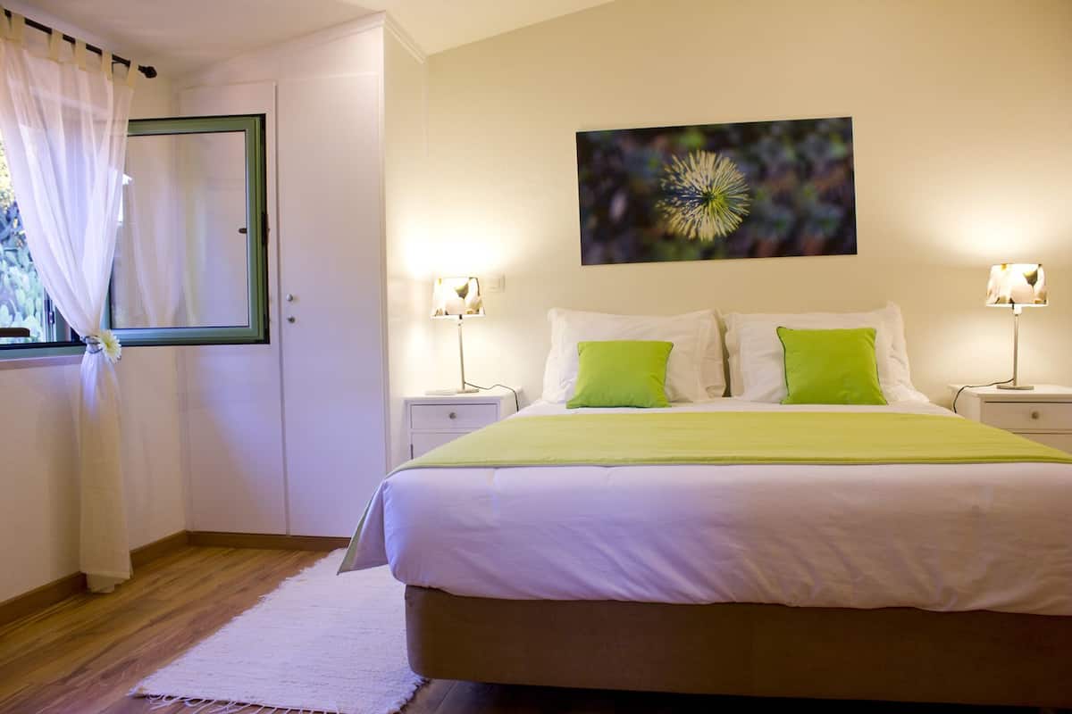 Quarto bem organizado com uma cama grande, duas mesas de cabeceira com luminárias, uma janela com cortinas leves e uma obra de arte na parede.
