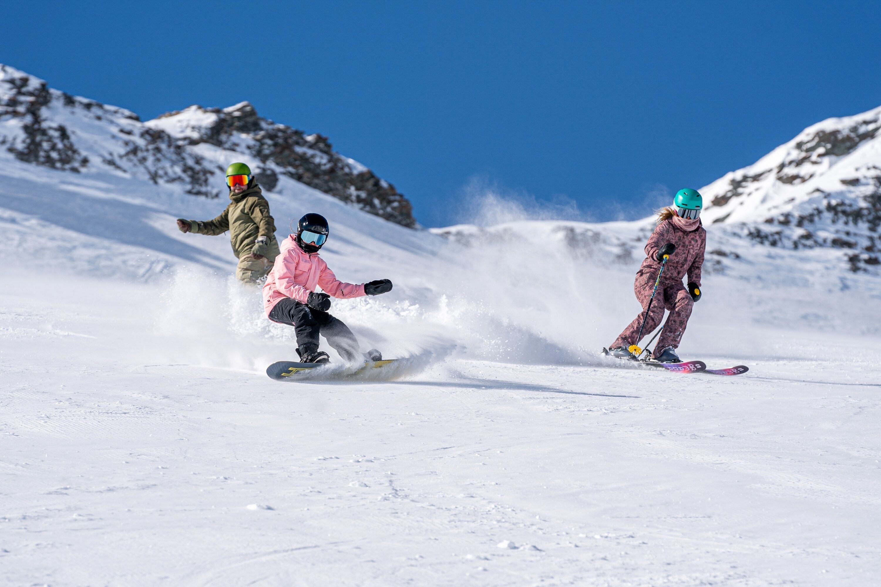Trois skieurs sur une pente enneigée, avec des montagnes en arrière-plan.