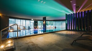 A modern indoor swimming pool with a glass barrier, sunlit windows, and a view of greenery outside. - Peaks Place (Laax)