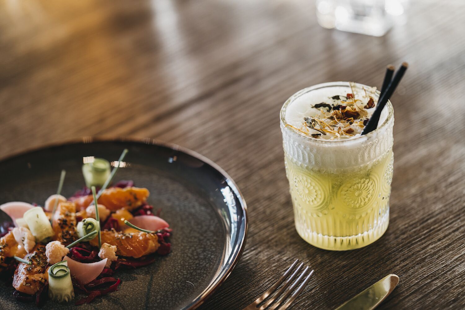 A wooden table with a plate of food and a glass of a creamy beverage with a straw.