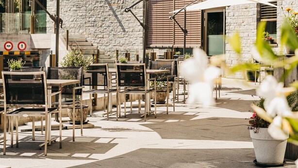 An outdoor cafe with tables and chairs under umbrellas, a stone building in the background, and a mountainous landscape. - Peaks Place (Laax)