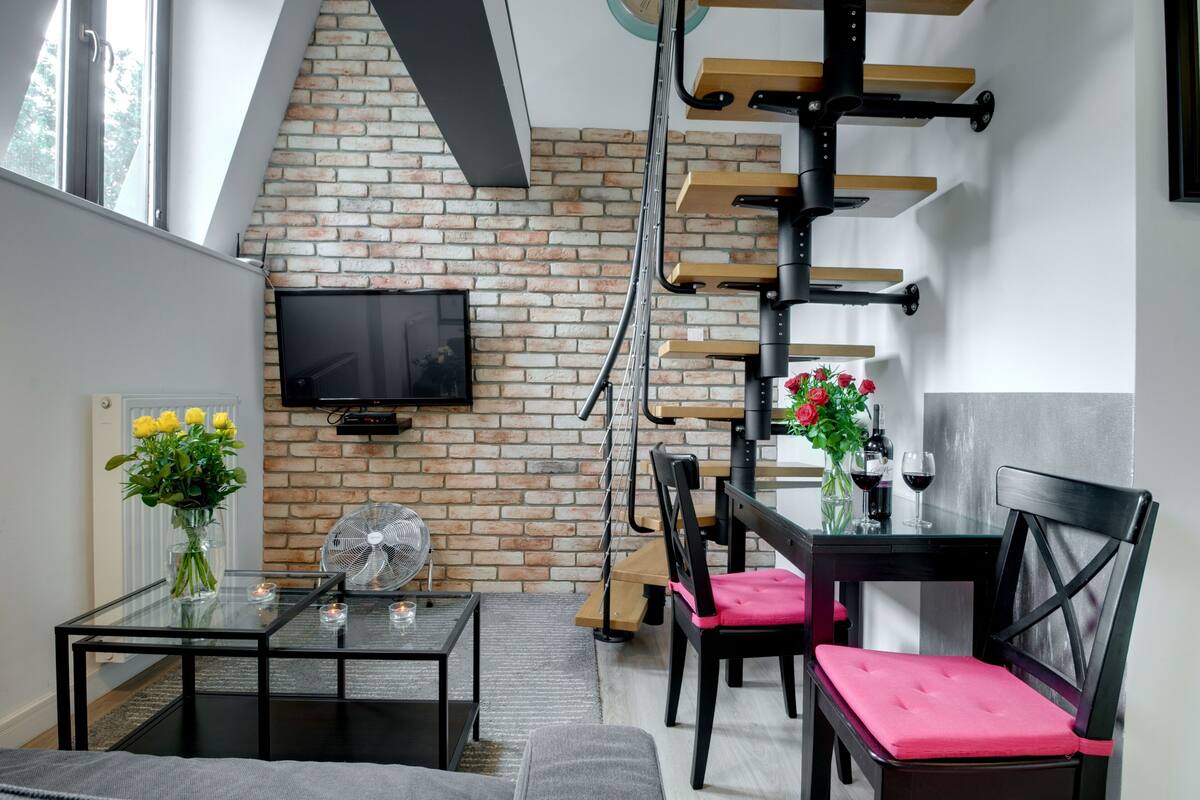 Een moderne woonkamer met een bakstenen muur, een aan de muur bevestigde televisie, een eettafel met stoelen en een trap met houten treden.