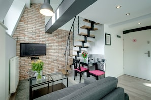 Un salon moderne avec un mur en briques, un téléviseur à écran plat, un coin repas avec une table et des chaises, et un escalier doté d’étagères.