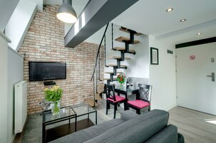 Egy modern nappali, téglából épített fallal, síkképernyős televízióval, étkezővel, melyben asztal és székek találhatók, valamint egyedi tervezésű lépcsővel.