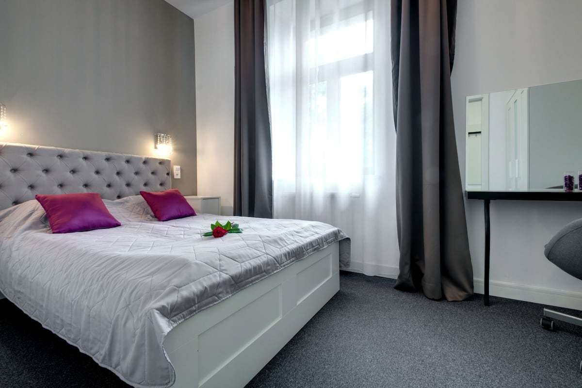 Een netjes ingerichte slaapkamer met een gestoffeerd hoofdeinde, een bed met een grijze quilt en twee roze kussens, een nachtkastje met een spiegel en een raam met dunne gordijnen.