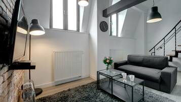 A modern living room with a sofa, coffee table, and a TV mounted on a brick wall. There are large windows and a staircase in the background.