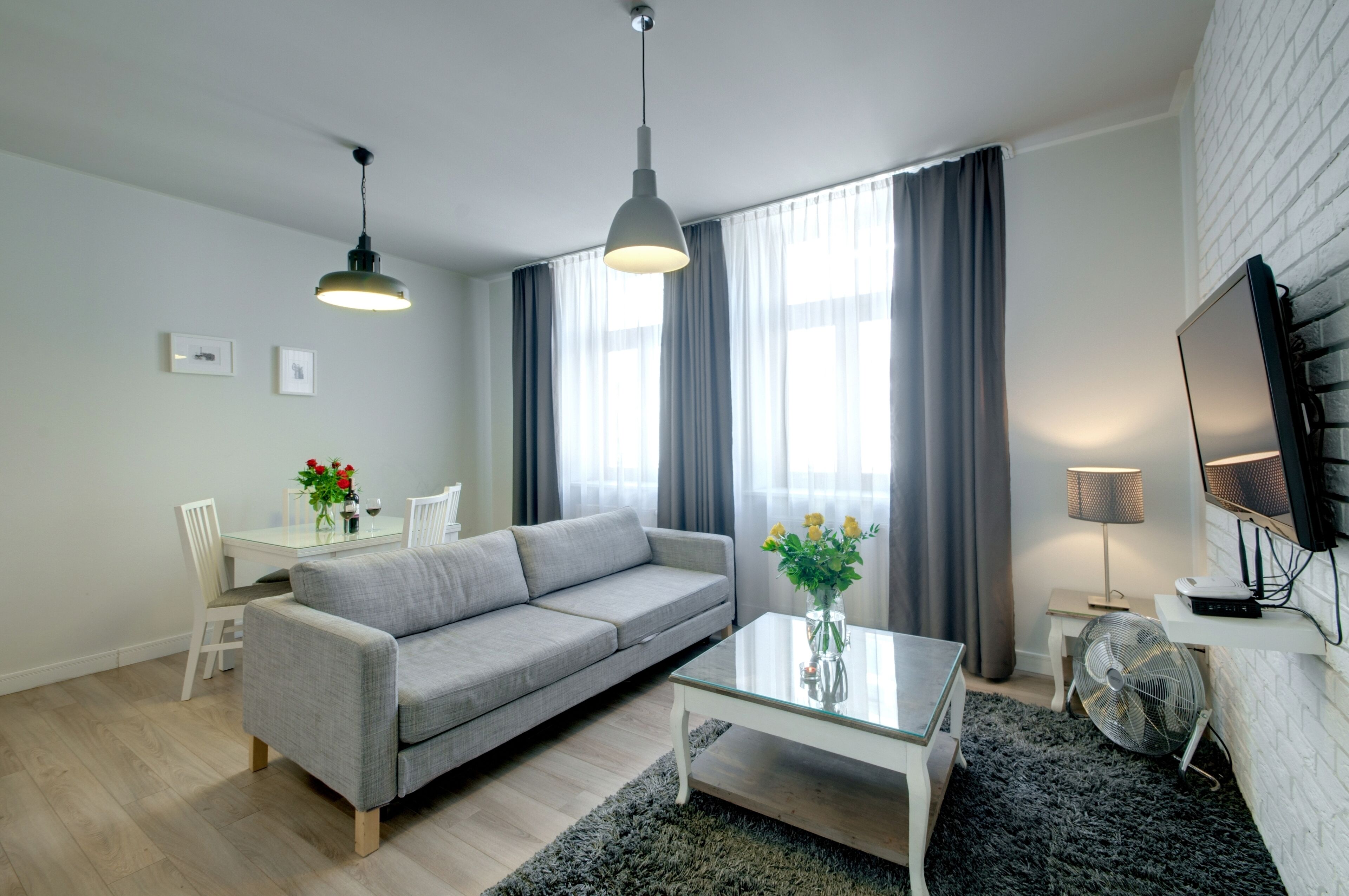 A modern living room with a grey sofa, a glass coffee table, a white TV stand, and a large window with grey curtains.