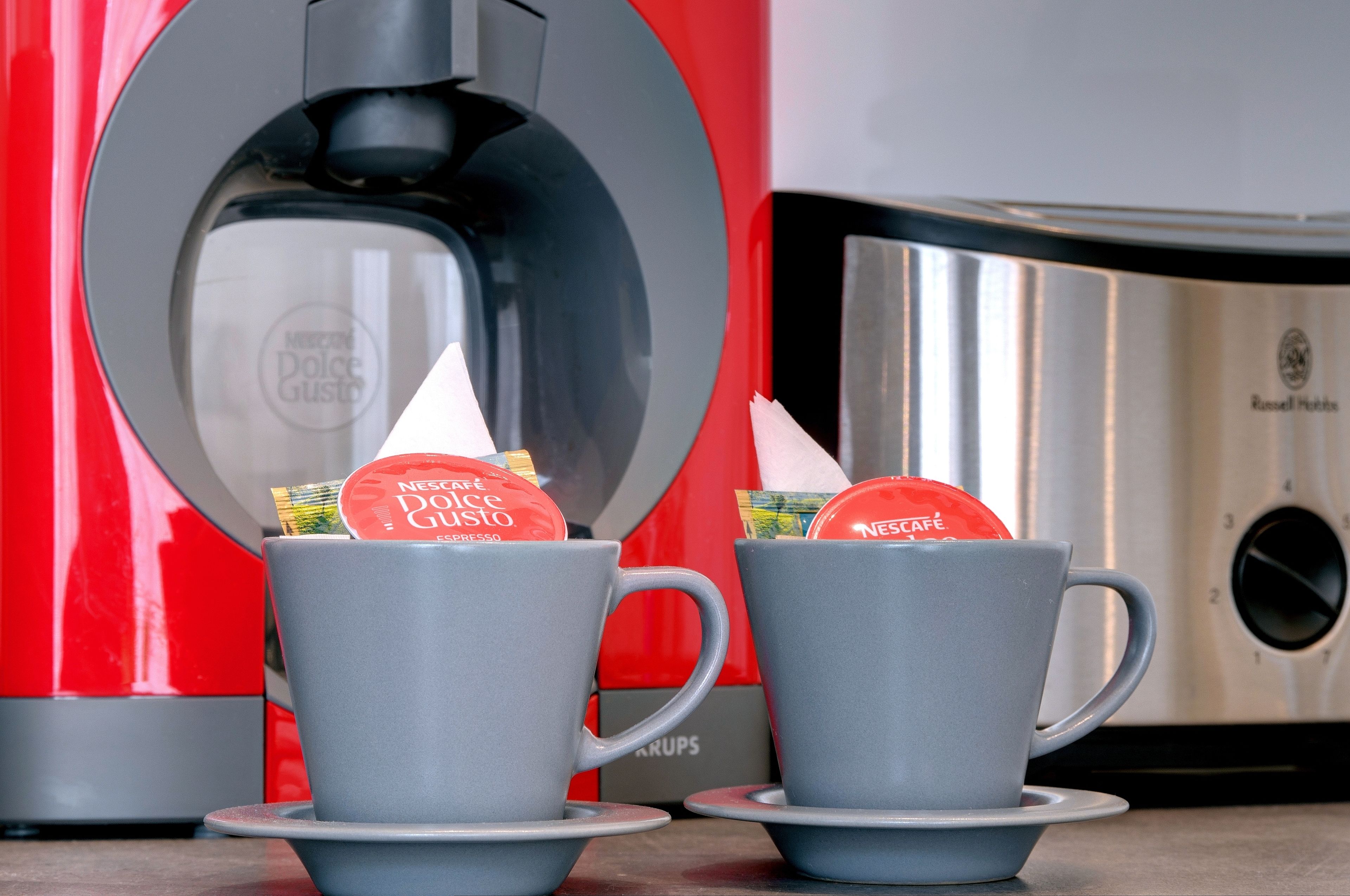 Two gray cups with Nespresso Dolce Gusto capsules on a table, a red and silver coffee machine, and a branded coffee pod box.