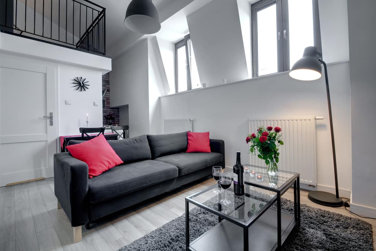 Een moderne woonkamer met een grijze bank, rode kussens, een glazen salontafel met wijnglazen en een fles, een vloerlamp, en uitzicht op een keuken en een raam.