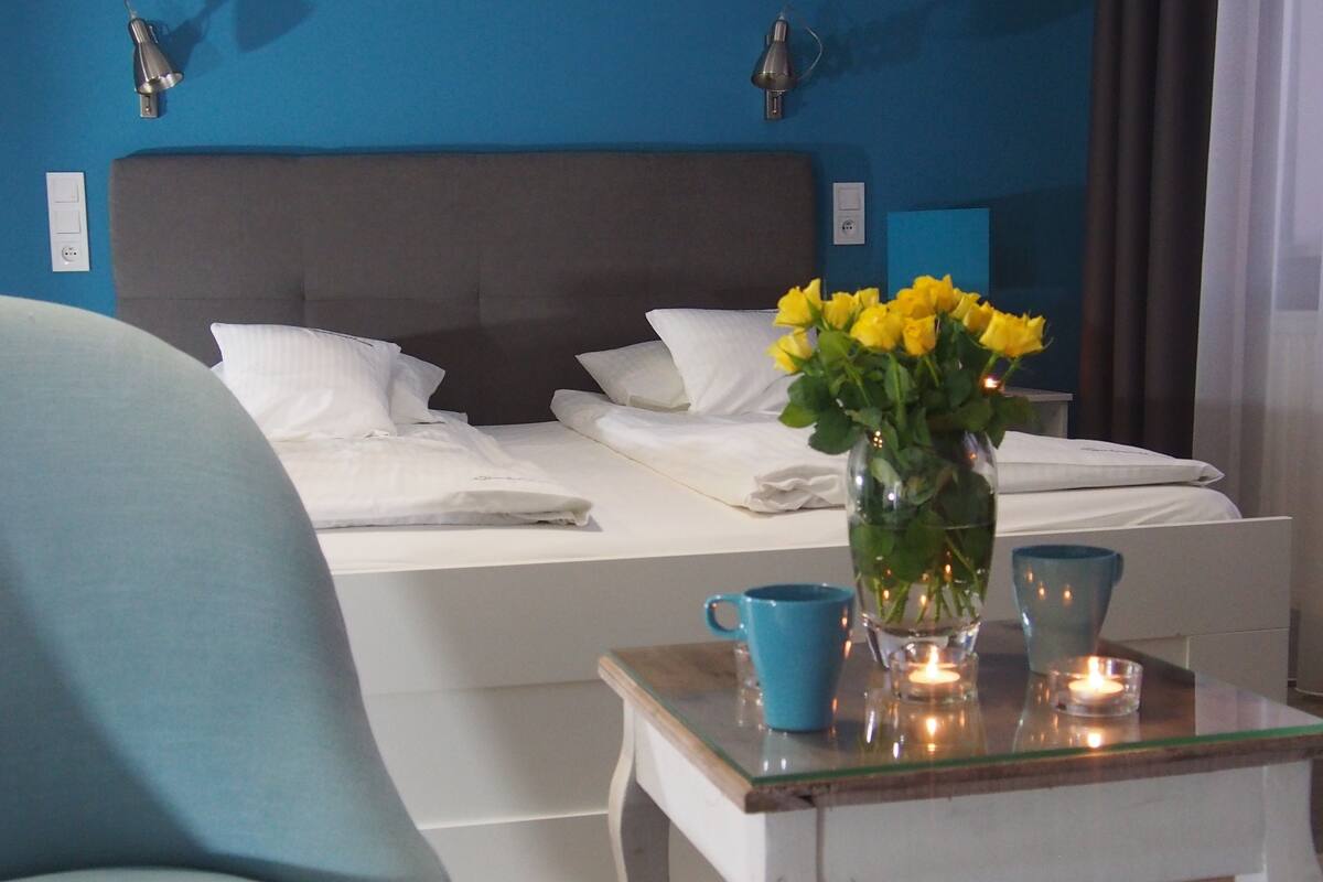 Een net opgemaakt bed met witte lakens, een vaas met gele bloemen, een blauwe mok en een brandende kaars op een bijzettafeltje.