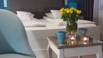 Un lit bien fait, avec du linge de lit blanc, un vase rempli de fleurs jaunes, une tasse bleue et une bougie allumée sur une petite table.