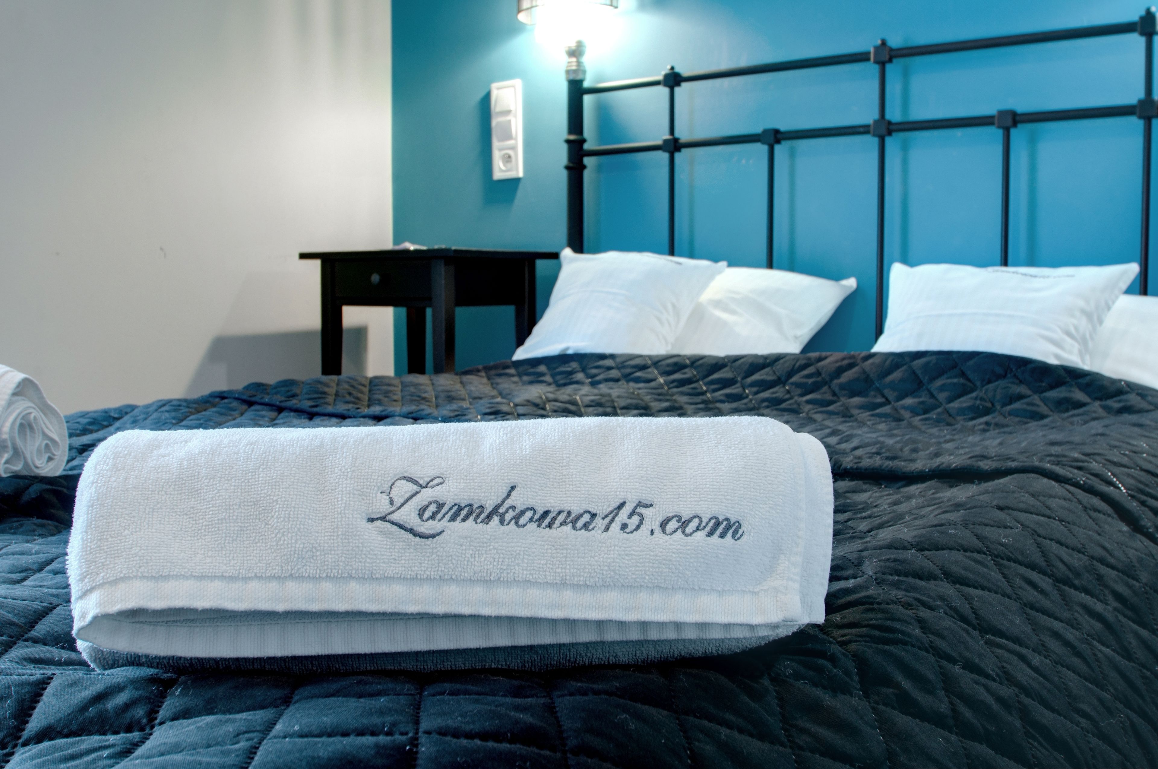 Un letto rifatto con un copriletto blu scuro e cuscini bianchi, un comodino e un portalampada fissato al muro.