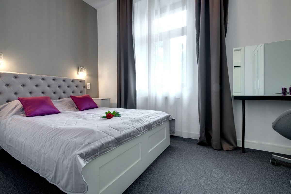 Een netjes ingerichte slaapkamer met een gestoffeerd hoofdeinde, een bed met een grijze quilt en twee roze kussens, een nachtkastje met een spiegel en een raam met dunne gordijnen.