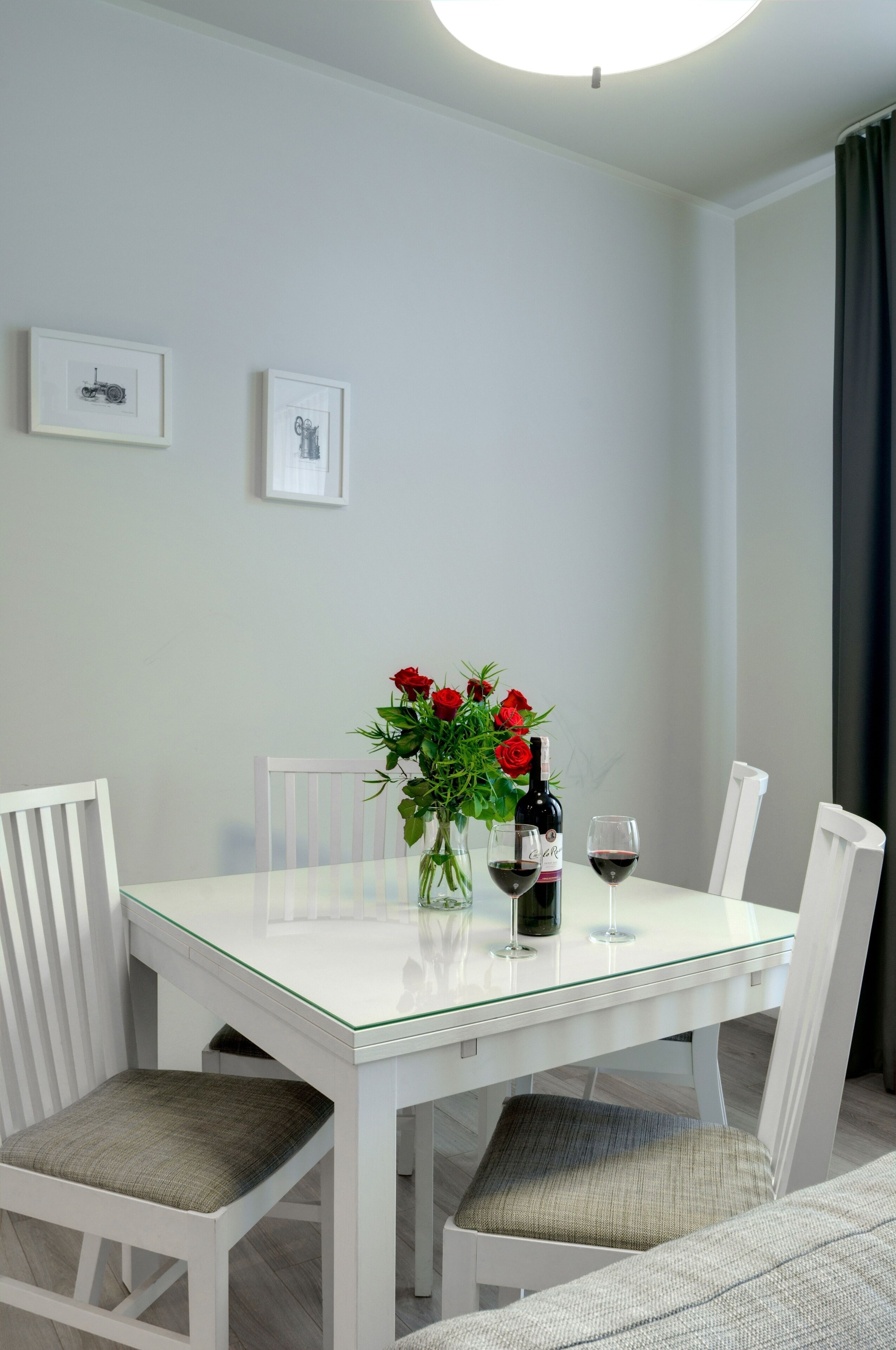 Een eetgedeelte met een glazen tafel, witte stoelen, een vaas rode rozen, een fles wijn en twee wijnglazen.