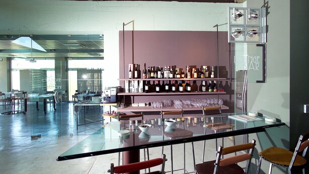 Un área de bar moderna con una mesa de vidrio, taburetes y una estantería con varias botellas de vino.