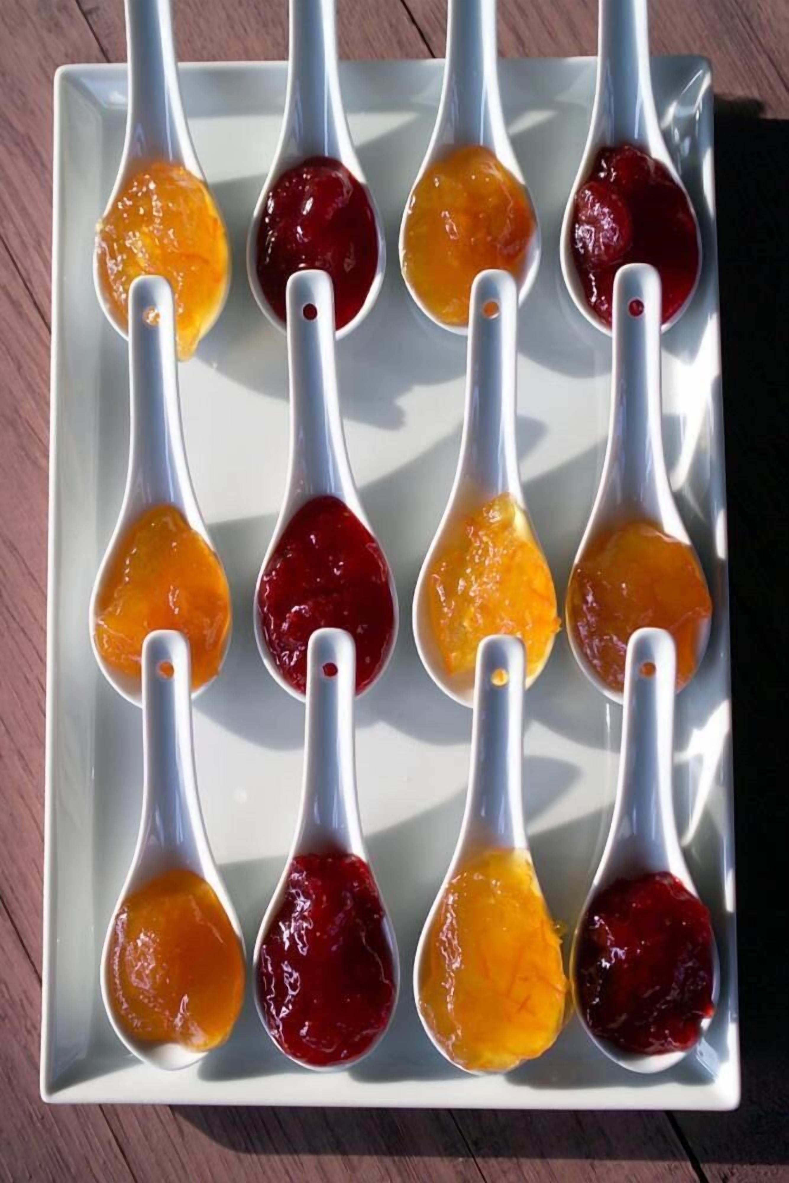 Un plato rectangular blanco con seis cucharas blancas llenas de jalea naranja y roja.