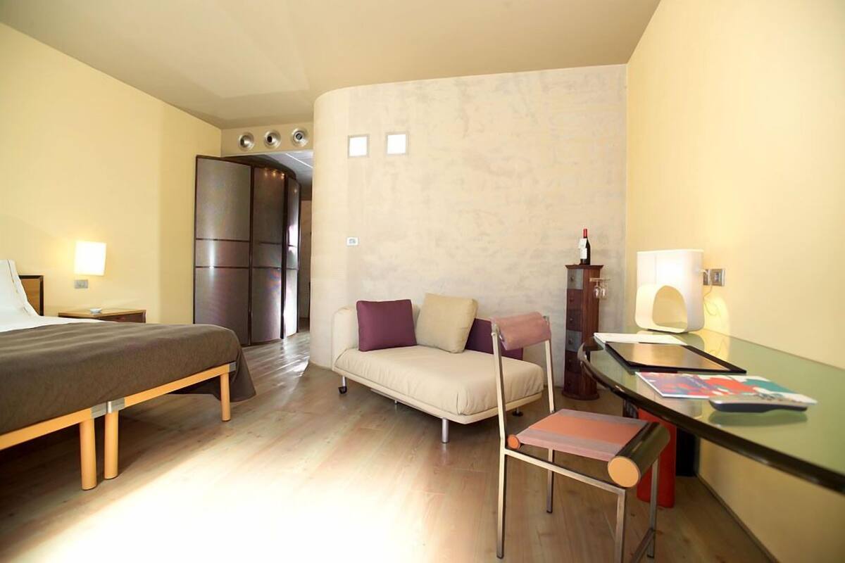 A hotel room with a bed, a sofa, a desk with a chair, a minibar, and a wall-mounted TV.