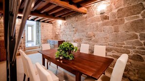 Un comedor con mesa de madera, sillas blancas, pared de piedra y techo de madera.