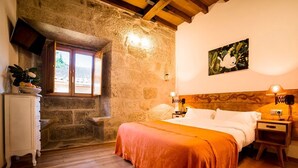 Un dormitorio con pared de piedra, suelo de madera, una cama con manta roja, una mesita de noche, una lámpara y un cuadro en la pared.