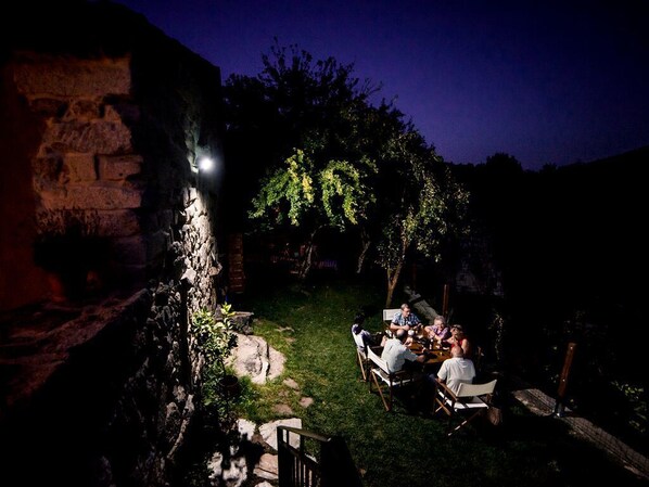 Un grupo de personas sentadas alrededor de una mesa al aire libre por la noche, con un muro de piedra y árboles de fondo.