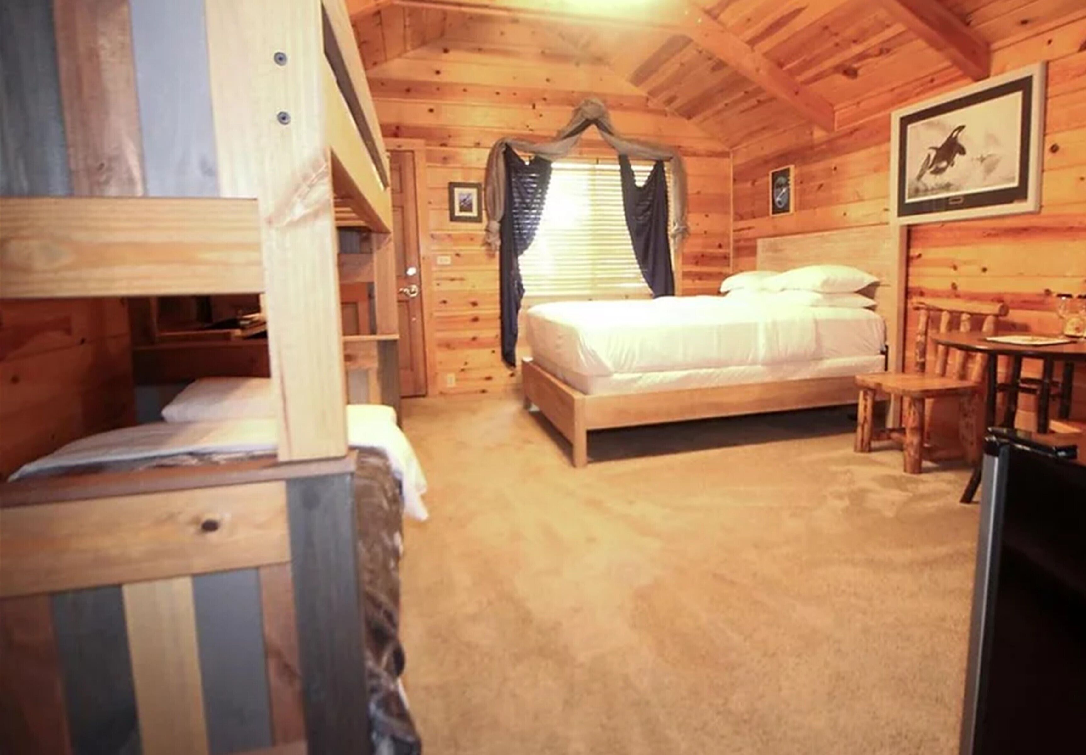 A wooden cabin room with a bunk bed, a single bed, a small table, and a framed picture on the wall.