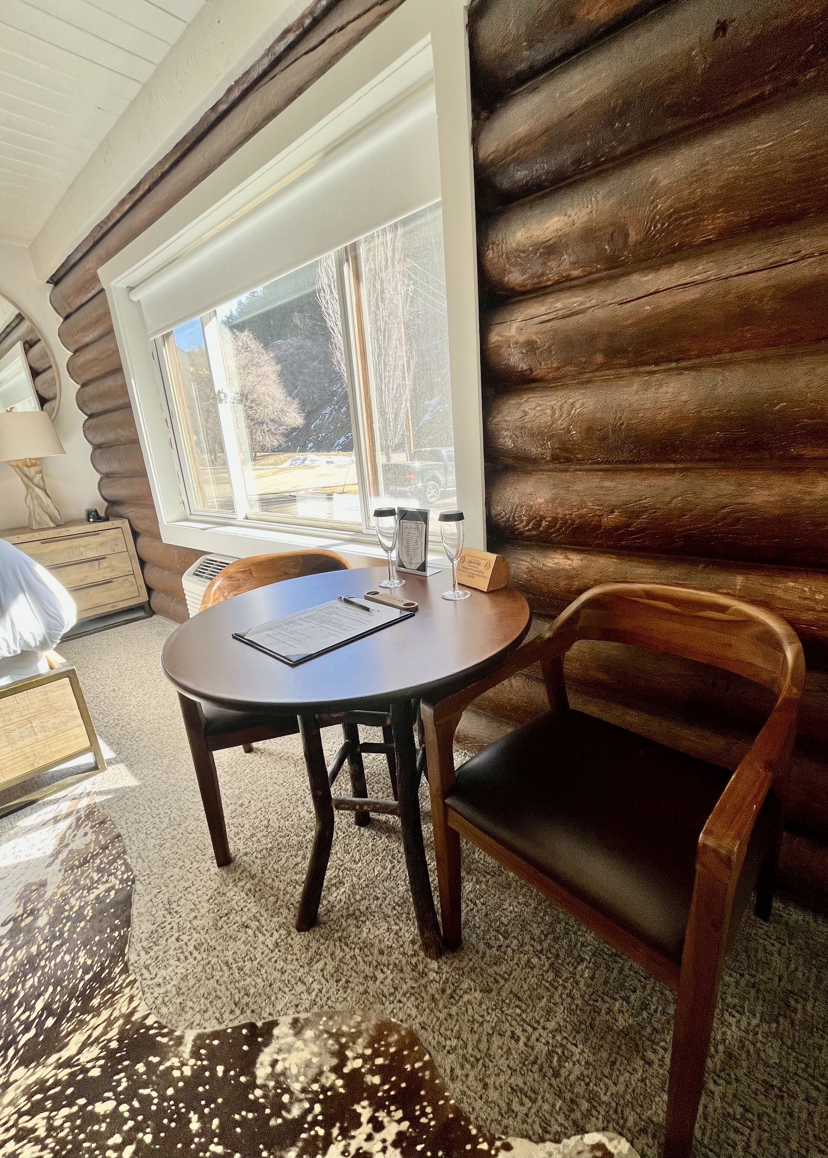 L’intérieur d’une cabane en rondins, avec une table ronde, deux chaises, une fenêtre donnant sur des arbres et un véhicule, ainsi qu’une lampe posée sur une commode.