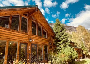 A log cabin with large windows, a staircase leading to the entrance, and a mountain backdrop.