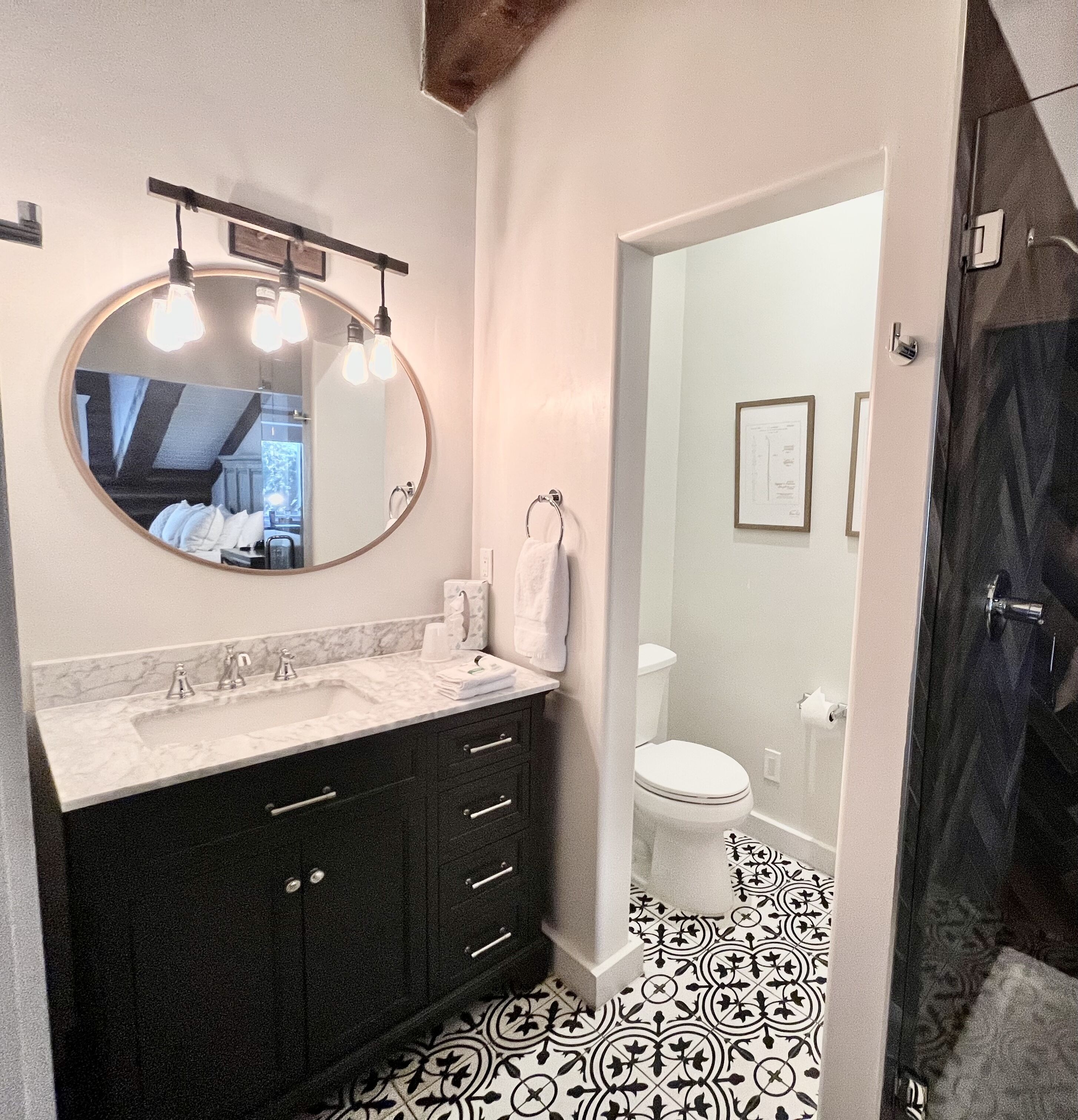 A bathroom with a round mirror, a sink with a marble countertop, dark cabinets, and a patterned floor.