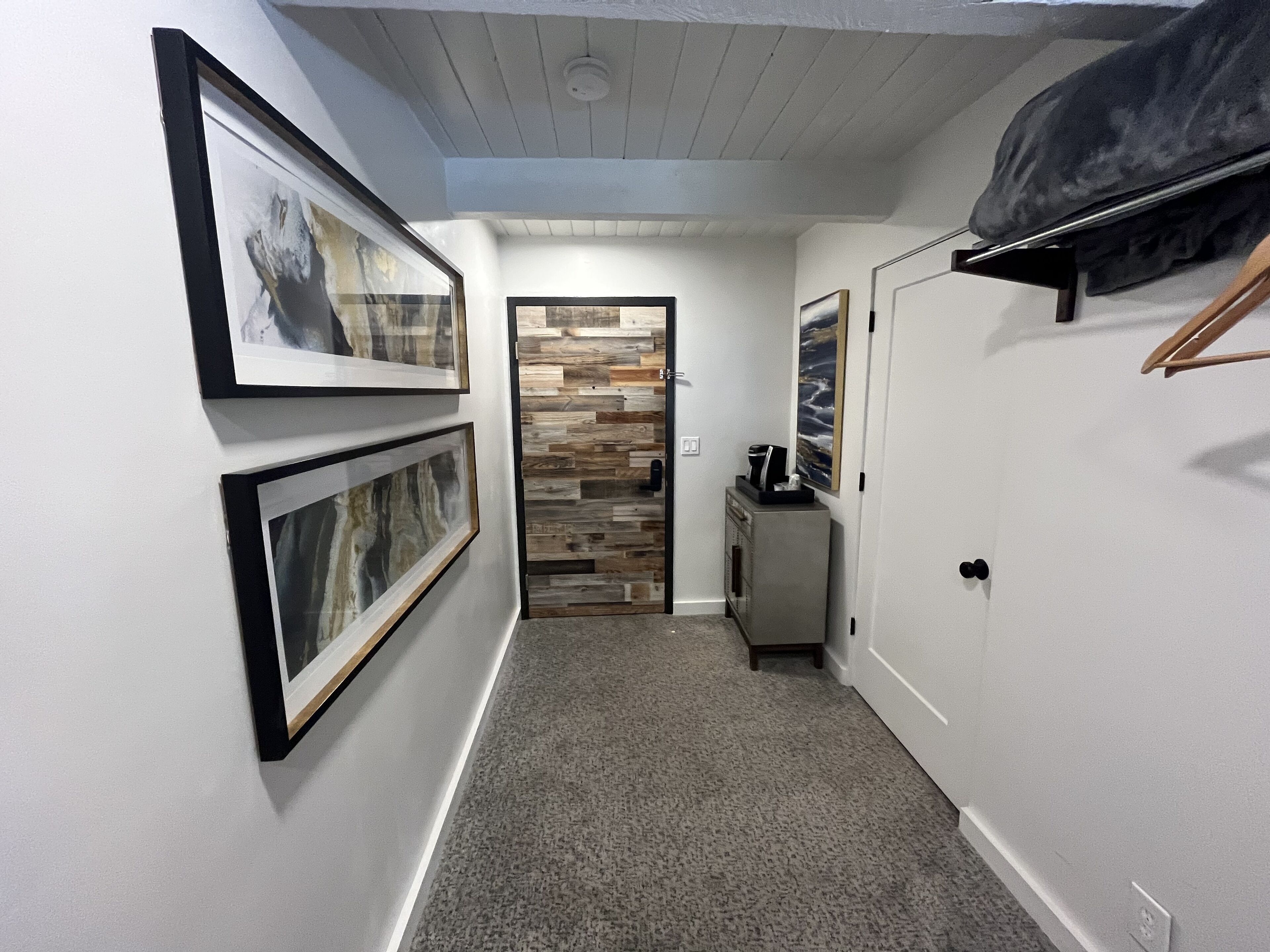 A hallway with framed artwork on the walls, a wooden door, and a small cabinet with a coffee maker.