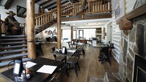A spacious dining area with wooden tables and chairs, a staircase, and a stone fireplace. - Alaskan Inn (Ogden)