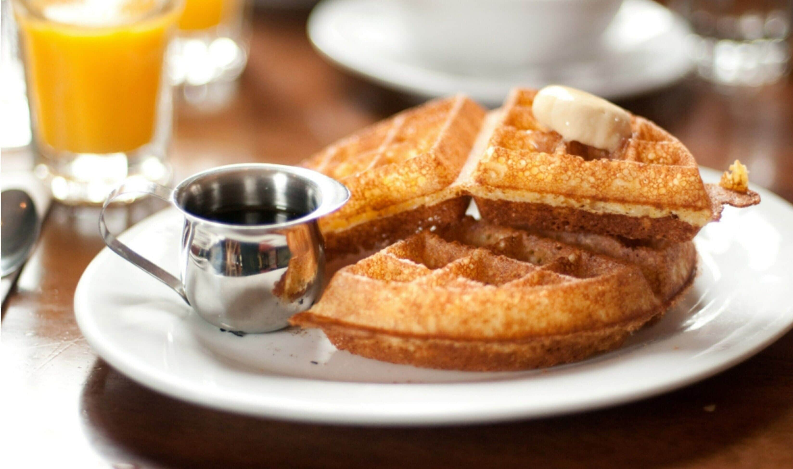 A plate of waffles with syrup and a pat of butter, accompanied by a small metal cup of syrup.
