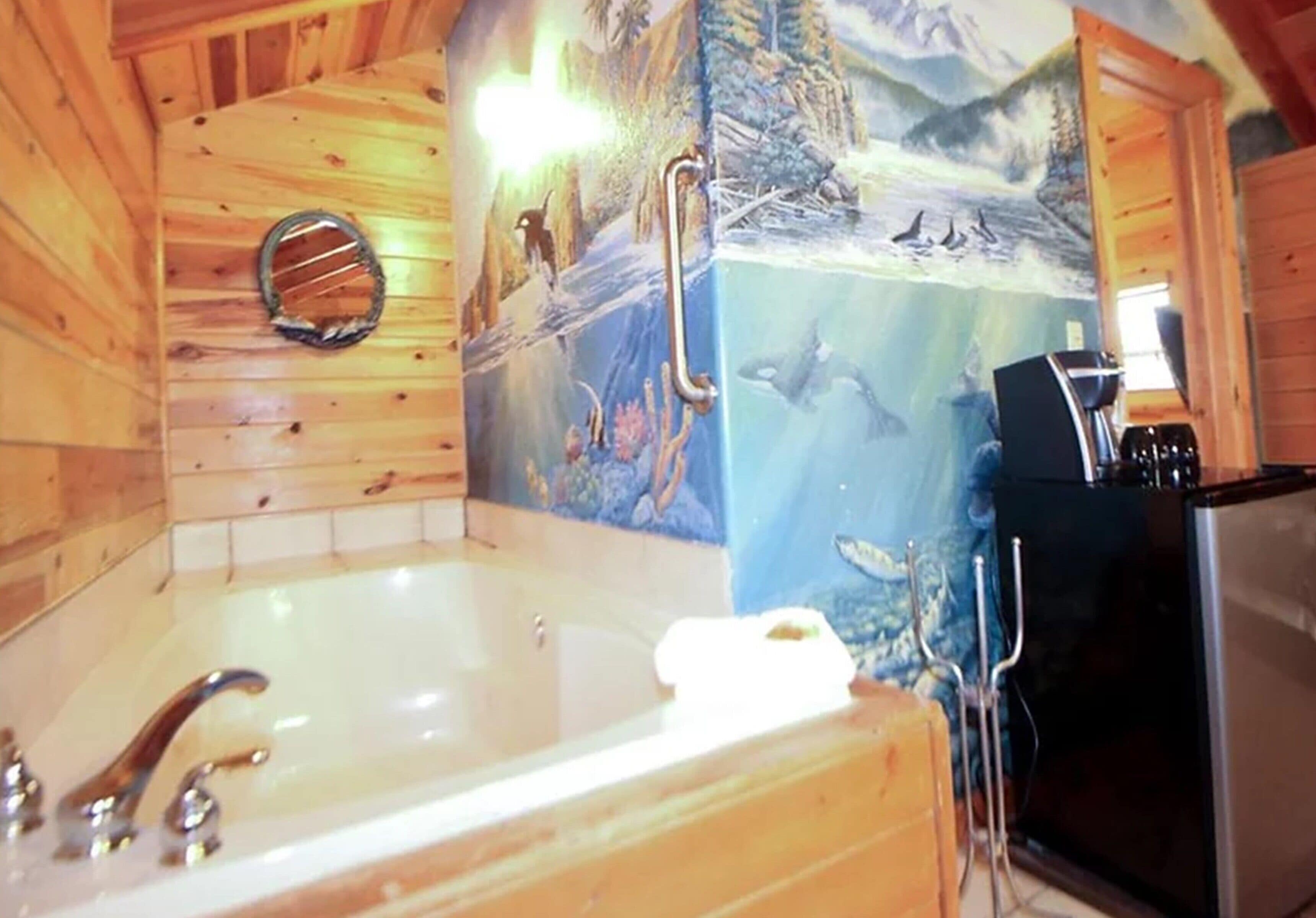 Interior de una cabaña de madera con una bañera grande, un mural de un paisaje montañoso y un refrigerador negro.