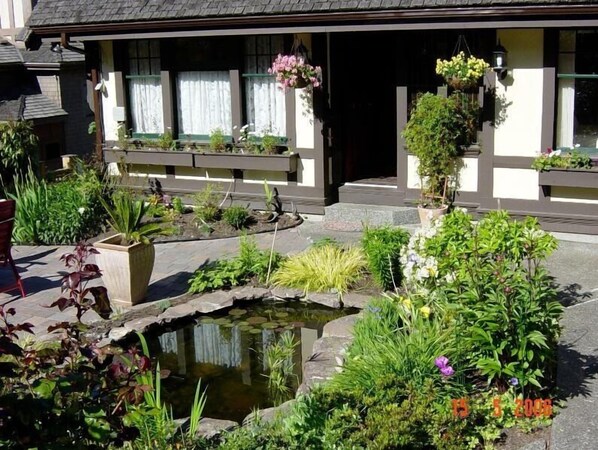 A garden with a pond, various plants, and a building with windows and hanging flowers.